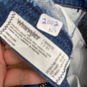 Pu&ograve; includere: Primo piano di un'etichetta di jeans Wrangler in denim blu con testo bianco. L'etichetta dice "Machine wash with like colors, tumble dry low, bleach when needed, iron medium, permanent press". L'etichetta dice anche "100% Cotton" e "153966 10-5E LIN9-02". C'&egrave; un'etichetta bianca con il testo "2002 NS" sul denim.