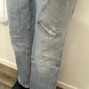 May include: Light blue denim jeans with distressed details and fading. The jeans have several small holes and tears on the front of the right leg.