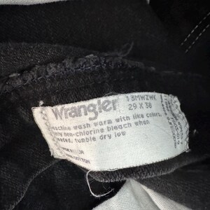 May include: Close-up of a black Wrangler jeans tag. The tag is white with black text, including the brand name and washing instructions. The jeans appear to be a dark wash, with visible stitching and a partially visible white pocket lining.