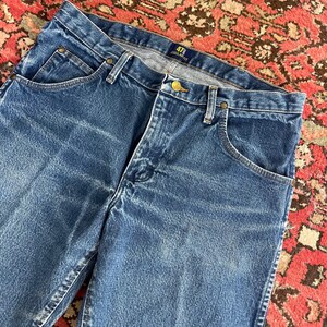 以下が含まれることがあります： クラシックな5ポケットデザインの青いデニムジーンズのクローズアップ。ジーンズはボタンとジッパーで開閉し、ベルトループがあり、ラベルには「47 Regular Fit」と書かれています。デニムは色あせ、使い古された外観をしています。