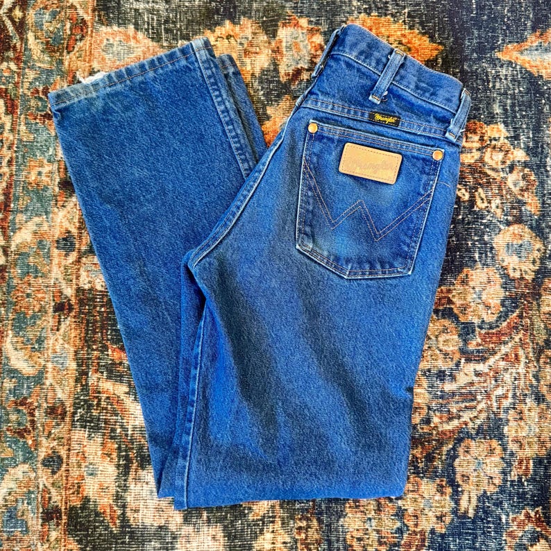 May include: Blue denim jeans with a classic straight leg cut. The jeans feature a Wrangler brand patch on the back pocket and a small logo above the pocket. The jeans are folded and laid flat.