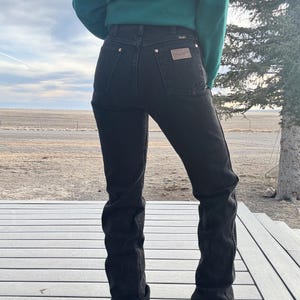 May include: Black denim jeans with a high waist and a classic five-pocket design. The jeans feature brass-colored rivets and a brown leather patch on the back pocket. The person is wearing a green sweater.