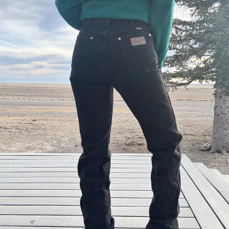 May include: Black denim jeans with a high waist and a classic five-pocket design. The jeans feature brass-colored rivets and a brown leather patch on the back pocket. The person is wearing a green sweater.