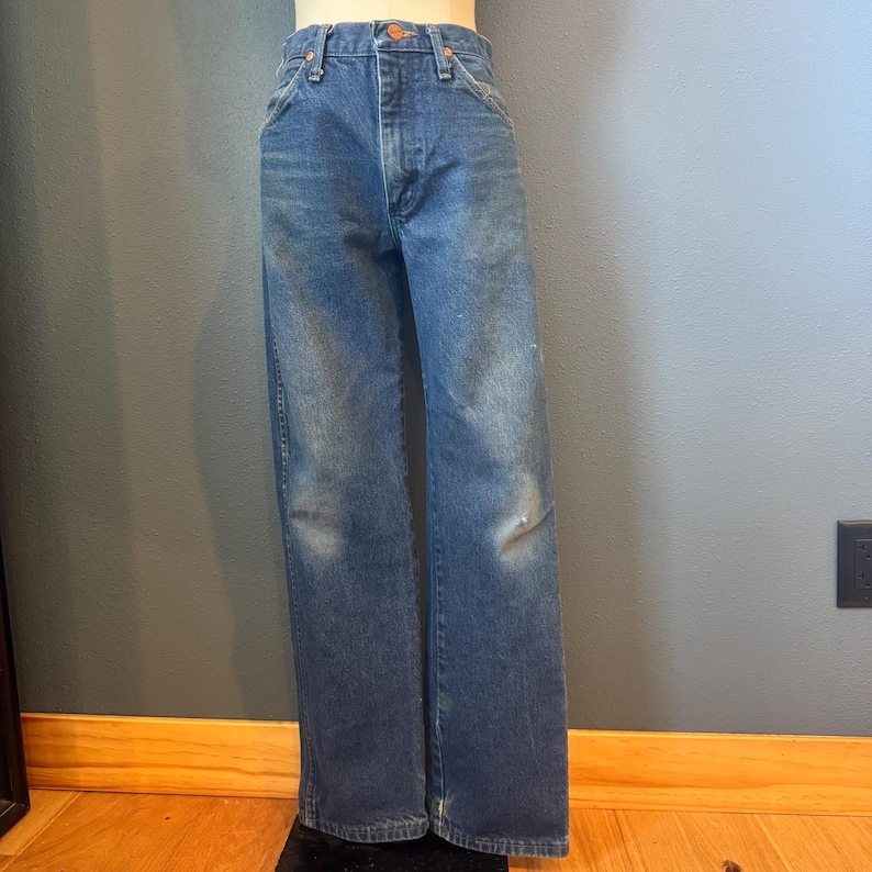 May include: Blue denim jeans with a classic straight leg fit. The jeans have a faded wash and show signs of wear, including light distressing on the knees. They feature a button and zipper closure, along with classic five-pocket styling.