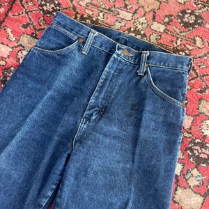 May include: Dark blue denim jeans with classic five-pocket styling. The jeans have a button and zipper closure, belt loops, and copper-colored rivets. The fabric shows signs of wear, adding to the vintage aesthetic. The jeans are folded.