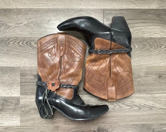 May include: A pair of brown and black cowboy boots with intricate stitching and a braided strap detail. The boots have a pointed toe and a low heel.