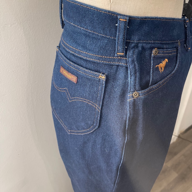 May include: Dark blue denim jeans with a brown leather patch that says "Wrangler" on the back pocket. The jeans have a horse logo on the right back pocket.