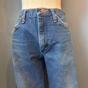 May include: Close-up of blue denim jeans with orange stitching and a copper-colored button. The jeans have a classic five-pocket design and a straight leg fit. The Wrangler brand name is visible on the button.