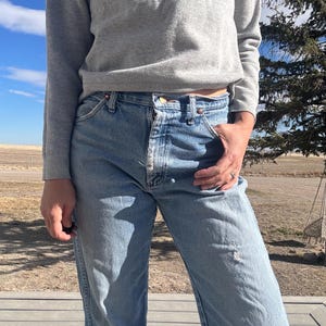 May include: Light blue denim jeans with a button fly and a distressed knee. The jeans are paired with a gray long-sleeved shirt. The person is standing outside on a wooden deck.