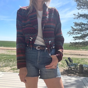 May include: A long-sleeve jacket featuring a multi-colored geometric pattern in shades of red, teal, and black. The jacket has a notched collar, a white undershirt, blue denim shorts, and a brown leather belt with a silver buckle and accents.