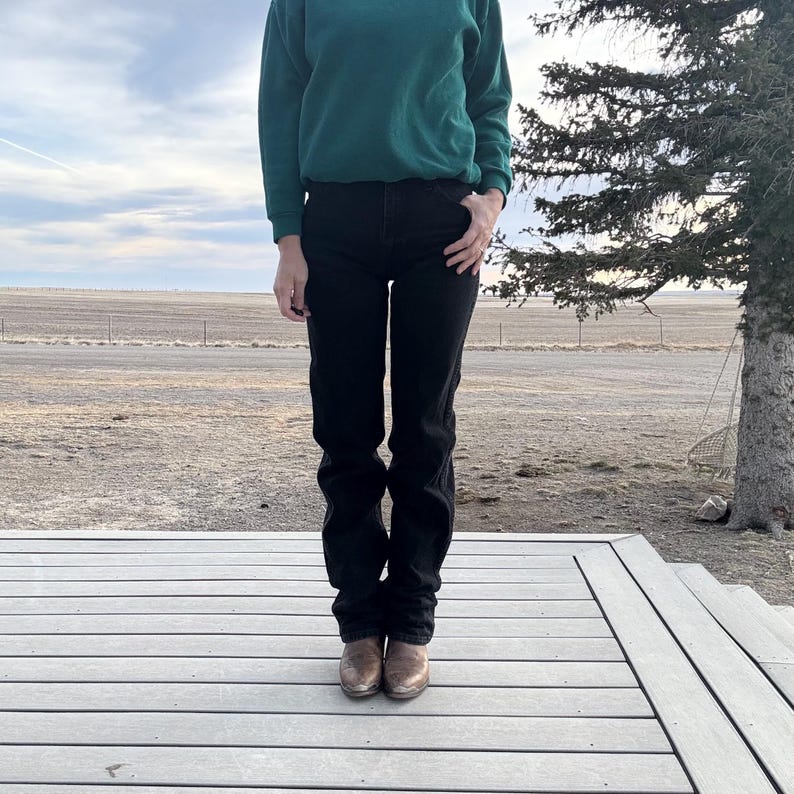 May include: A person wearing a dark green sweatshirt, black jeans, and brown cowboy boots. The person is standing on a wooden deck with a rural landscape in the background. The jeans are straight-legged and the boots are worn.