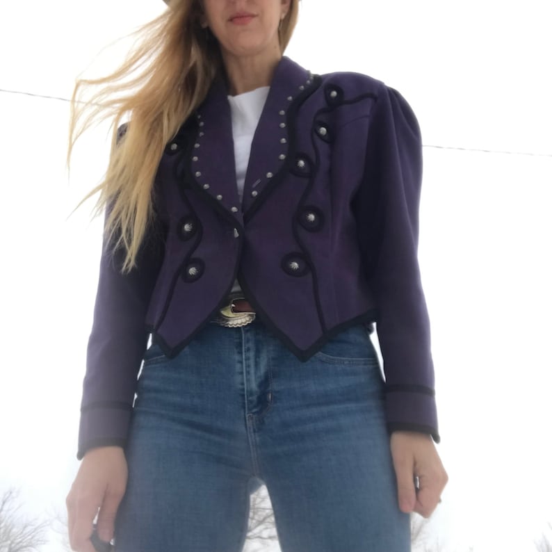 May include: A purple cropped jacket with black trim and silver accents. The jacket has a pointed collar and a double-breasted closure.