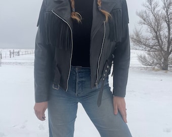 May include: A black leather jacket with fringe detailing. The jacket has a zipper closure and a belt at the waist. The model is wearing blue jeans.