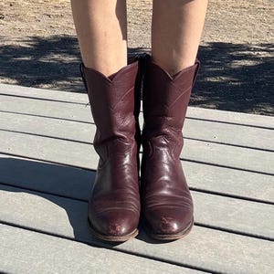 May include: A pair of burgundy leather cowboy boots. The boots have a classic Western design with pointed toes and angled heels. The leather shows signs of wear, adding to their vintage appeal.