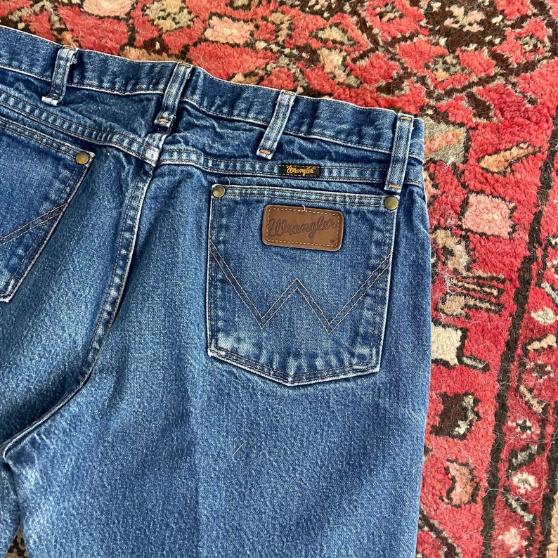以下が含まれることがあります： バックポケットにWranglerのラベルが付いた、青いデニムジーンズのクローズアップ。 ジーンズは、ボタンとジッパー留めを備えたクラシックな5ポケットデザインです。 ステッチは対照的なタン色です。