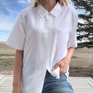 May include: A short-sleeved white button-down shirt featuring a pointed collar with delicate white embroidery, a central vertical panel with fine pleats, and intricate floral embroidery running down the center front placket of the garment.