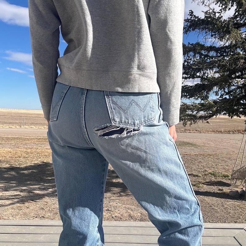 May include: Light blue denim jeans with a distressed tear on the back pocket. The jeans have a classic five-pocket design and a straight leg fit. The person is wearing a gray sweatshirt.