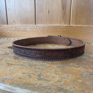May include: A brown leather belt with a floral design. The belt is embossed with a repeating pattern of leaves and flowers.