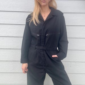 May include: A black long-sleeved jumpsuit with a collared neckline and a belt. The jumpsuit has two zippered pockets on the chest and two zippered pockets on the hips.