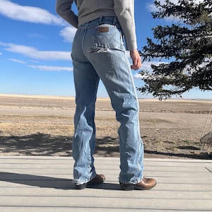 May include: Light blue denim jeans with a distressed back pocket. The jeans have a classic fit and are paired with brown cowboy boots. The person is wearing a gray sweater. The background features a blue sky and a rural landscape.