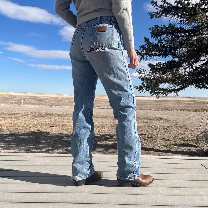 May include: Light blue denim jeans with a distressed back pocket. The jeans have a classic fit and are paired with brown cowboy boots. The person is wearing a gray sweater. The background features a blue sky and a rural landscape.