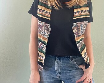 May include: A black sleeveless vest with a colourful geometric pattern. The vest has a V-neck and is made of a woven fabric. The vest is being worn over a black t-shirt and blue jeans.