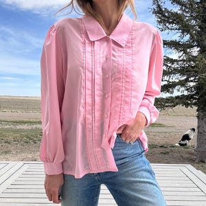 May include: A light pink button-down blouse with long sleeves and a classic collar. The blouse features vertical pleats down the front and is paired with blue jeans. The sleeves have buttoned cuffs.