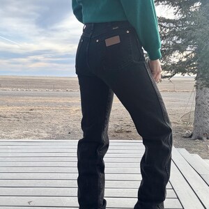 May include: Black denim jeans with a high waist and a straight leg. The jeans have a brown leather patch on the back pocket and gold-colored rivets. The person is wearing a green sweater. The jeans are a classic style.