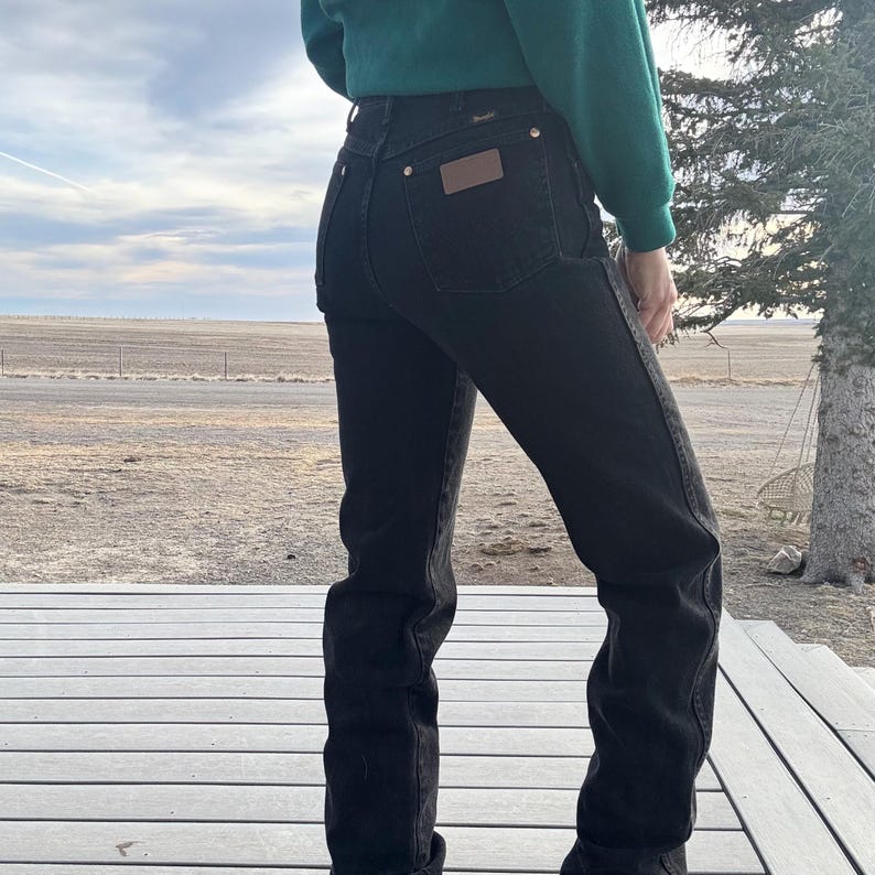 May include: Black denim jeans with a high waist and a straight leg. The jeans have a brown leather patch on the back pocket and gold-colored rivets. The person is wearing a green sweater. The jeans are a classic style.