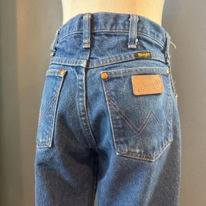 May include: Close-up of vintage blue denim Wrangler jeans. The jeans feature a classic five-pocket design, copper rivets, and a brown leather patch with the Wrangler logo. The back pockets have a signature 'W' stitching.