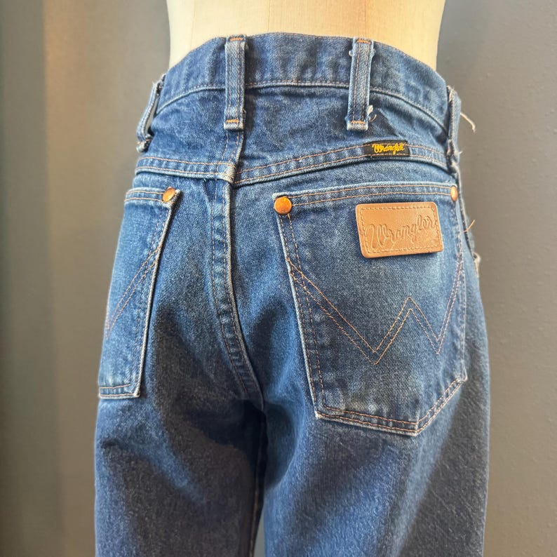 May include: Close-up of vintage blue denim Wrangler jeans. The jeans feature a classic five-pocket design, copper rivets, and a brown leather patch with the Wrangler logo. The back pockets have a signature 'W' stitching.
