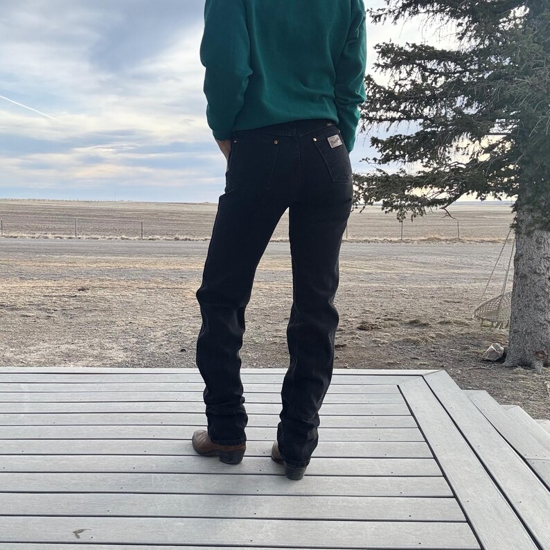 May include: A person wearing a dark green sweater and black jeans with brown cowboy boots. The person is standing on a wooden deck with a rural landscape in the background.
