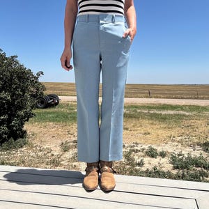 May include: Light blue trousers with a straight leg, featuring a classic design. The trousers have belt loops and pockets. The person is wearing brown suede boots. The background shows a clear blue sky and a field.