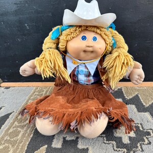 Op de afbeelding: Een pop met blond wollen haar in vlechten, met een witte cowboyhoed, een geruit overhemd met blauwe knopen en een bruine rok in suède-look met franjes. De pop heeft blauwe ogen en een lichte huidskleur, gekleed in een westernstijl.