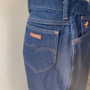 May include: Close-up of a pair of dark blue denim jeans with a brown leather patch that says "Wrangler" on the back pocket.