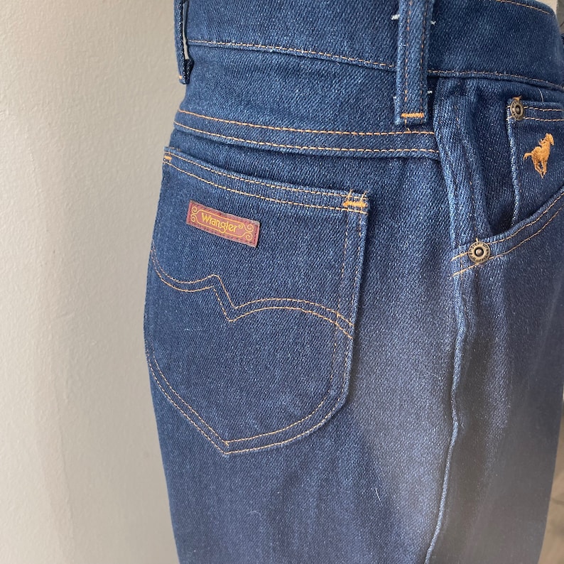 May include: Close-up of a pair of dark blue denim jeans with a brown leather patch that says "Wrangler" on the back pocket.