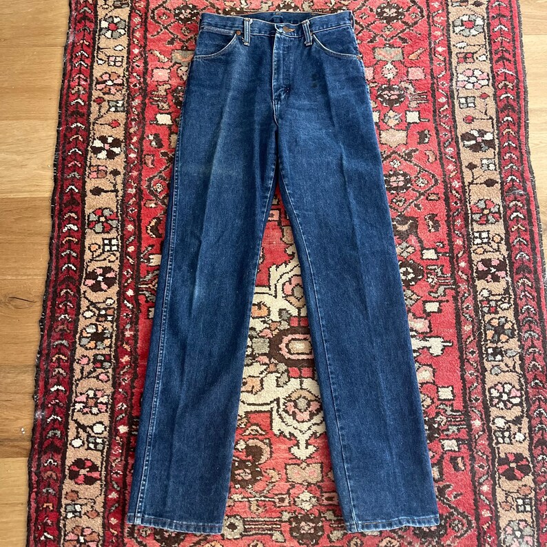 May include: Dark blue denim jeans with a high waist and straight leg. The jeans are laid flat on a patterned rug, showcasing the classic five-pocket design and button closure. The denim has subtle fading.