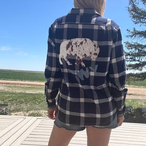 May include: A long-sleeve button-down shirt featuring a navy blue and white plaid pattern. The back of the shirt displays a large applique of a buffalo with a brown and white spotted hide texture, positioned above the blue embroidered letters WY.