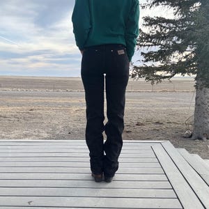 May include: Black denim jeans with a classic fit. The jeans have a straight leg and a dark wash. The person is wearing a green sweater. The background shows a cloudy sky and a wooden deck.