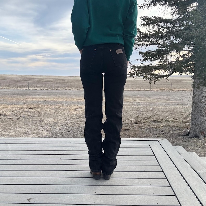 May include: Black denim jeans with a classic fit. The jeans have a straight leg and a dark wash. The person is wearing a green sweater. The background shows a cloudy sky and a wooden deck.