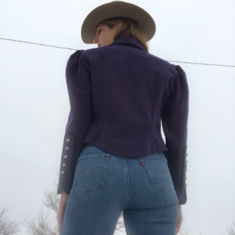May include: A purple cropped jacket with puffed shoulders and silver buttons on the cuffs. The jacket is worn over blue jeans.
