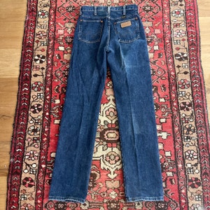 May include: Dark blue denim jeans with a classic five-pocket design. The jeans feature a brown leather patch on the back pocket and a Wrangler label. The jeans are laid flat on a patterned red and beige rug.