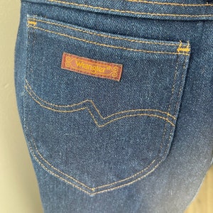 May include: A close-up of a dark blue denim back pocket with a brown leather Wrangler label. The pocket has a curved top and a stitched design.