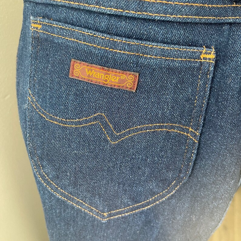 May include: A close-up of a dark blue denim back pocket with a brown leather Wrangler label. The pocket has a curved top and a stitched design.