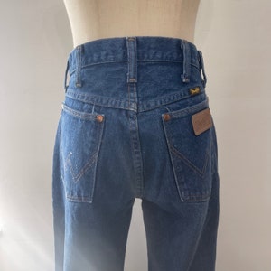 Pu&ograve; includere: Un paio di jeans blu denim con una toppa in pelle marrone sulla tasca posteriore che dice "Wrangler".