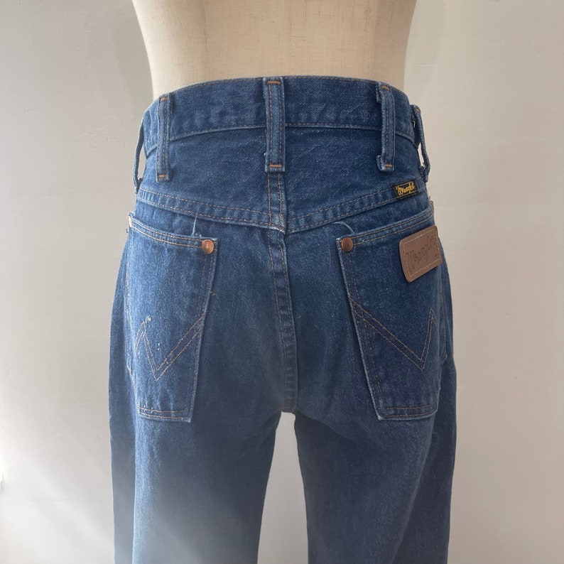 Pu&ograve; includere: Un paio di jeans blu denim con una toppa in pelle marrone sulla tasca posteriore che dice "Wrangler".