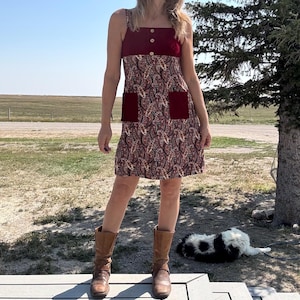 Può includere: Un vestito senza maniche con corpetto bordeaux e due bottoni in legno. L'abito presenta un motivo paisley nei toni del rosso, marrone e crema, con due tasche bordeaux. La modella indossa stivali marroni.
