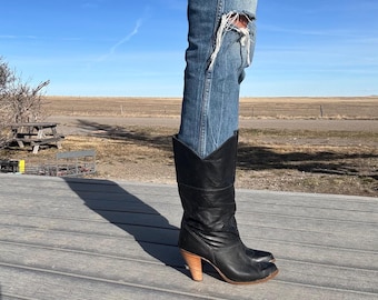 May include: Black leather cowboy boots with a stacked wooden heel. The boots have a pointed toe and a decorative stitched design. The boots are worn with blue denim jeans with a distressed knee.