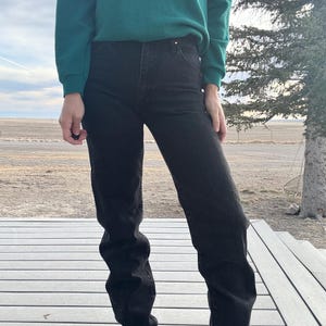 May include: Black denim jeans with a high waist. The jeans are paired with a dark green long-sleeved shirt. The person is standing on a wooden deck with a rural landscape in the background.