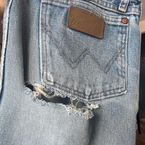 May include: Close-up of light blue denim jeans with a distressed, ripped design on the back pocket area. The jeans feature a brown leather Wrangler patch and a classic pocket design.